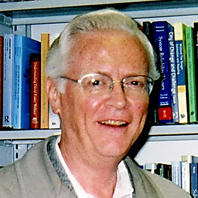 A man in a gray jacket, white shirt, and eyeglasses smiles at the camera.